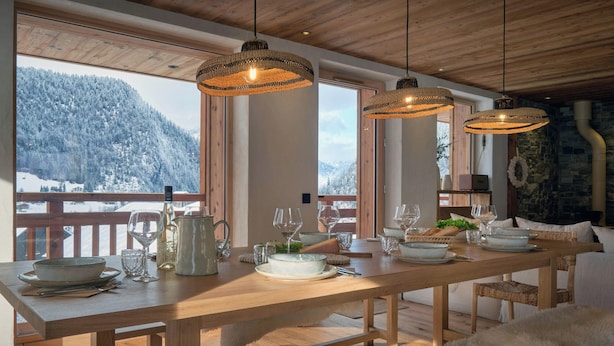 Una mesa de comedor de madera dispuesta con platos, vasos y una jarrón, con vistas a un paisaje montañoso nevado.