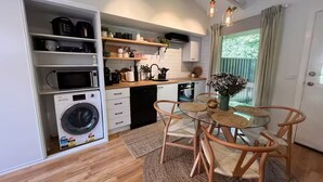Une cuisine compacte dotée d’appareils électroménagers intégrés, un coin repas avec une table en verre et des chaises en osier, et une vue sur l’extérieur par une fenêtre.