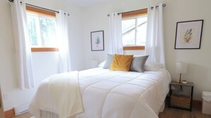 A bedroom with a large bed, white curtains, a bedside table with a lamp, and framed artwork on the walls. - Hare Hideaway with Fireplace (Prince Edward Island)