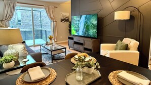 Un salon moderne doté d’un grand téléviseur à écran plat, d’une table à manger dressée avec des sets de table et un vase de fleurs, ainsi que d’un canapé agrémenté de coussins.