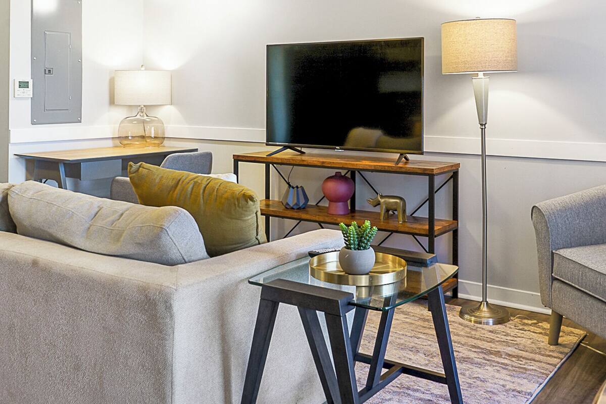 Una sala moderna con sofá, sillón, mesa de centro, televisor, lámpara y un estante pequeño con adornos.