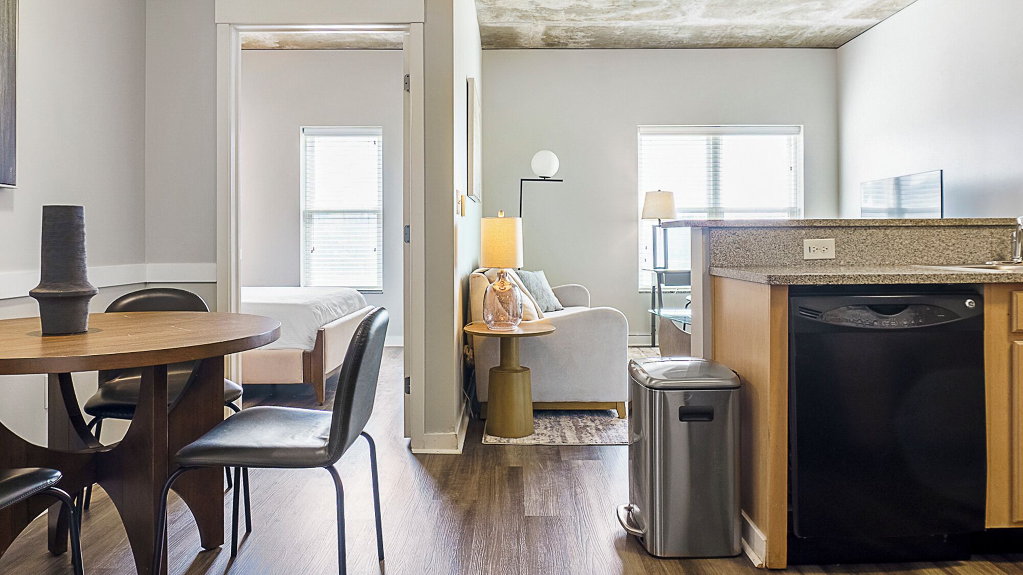 A modern kitchen with a dishwasher, a dining area with a table and chairs, and a living area with a sofa and a lamp.
