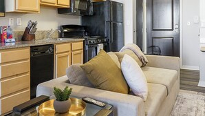 A compact living space with a kitchenette, a sofa with cushions, a small table with a plant and remote controls, and a door in the background. - Canal Walk District Apartments by Landing (Indianapolis)
