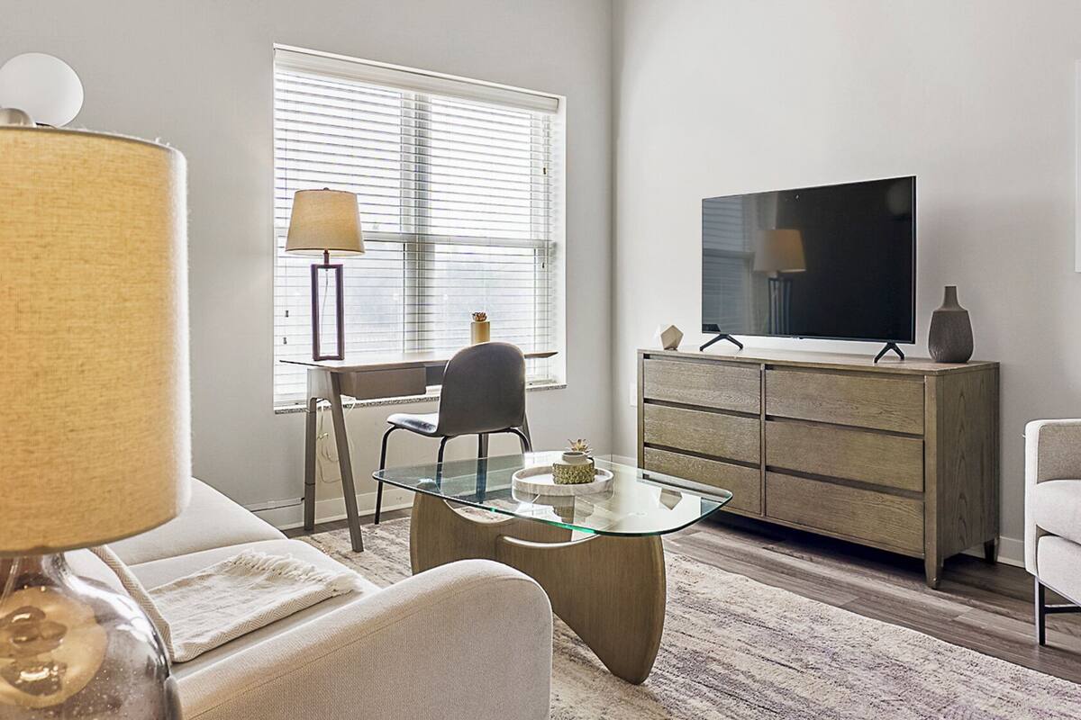 Una sala moderna con una mesa de centro de vidrio, un escritorio con una silla, un sofá, un televisor sobre un mueble de madera y una lámpara.