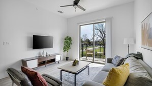 A modern living room with a flat-screen TV, a sofa, a coffee table, and a balcony with a view. - Landing Apartments Denton (Denton)
