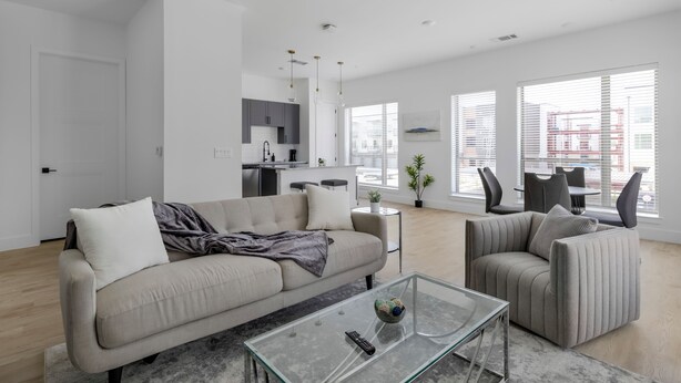 A modern living room with a sofa, armchair, and coffee table. There is a kitchen in the background with a dining area visible through large windows. - Landing Apartments Denton (Denton)