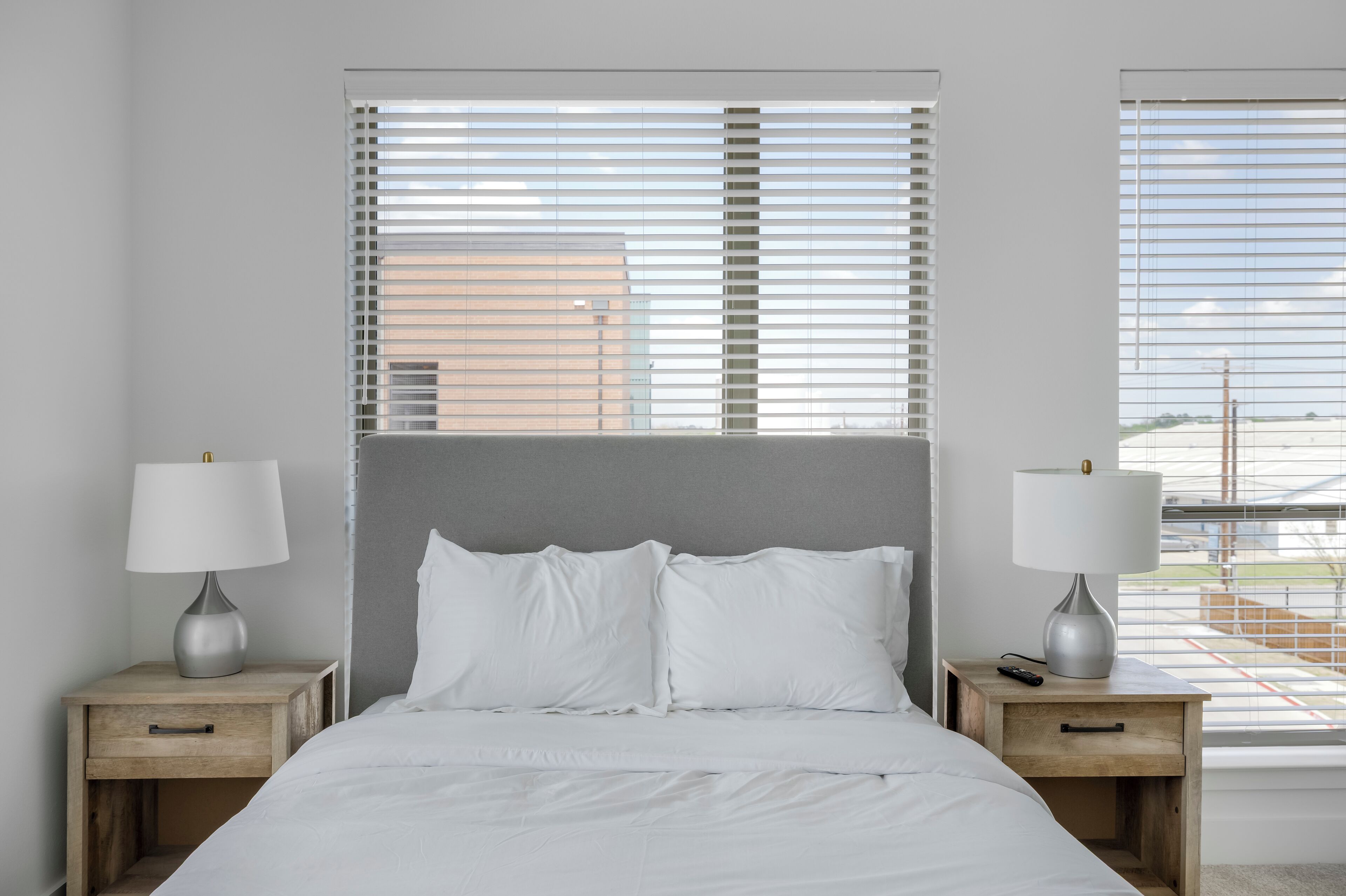 A bedroom with a bed, two bedside tables with lamps, and large windows with blinds.