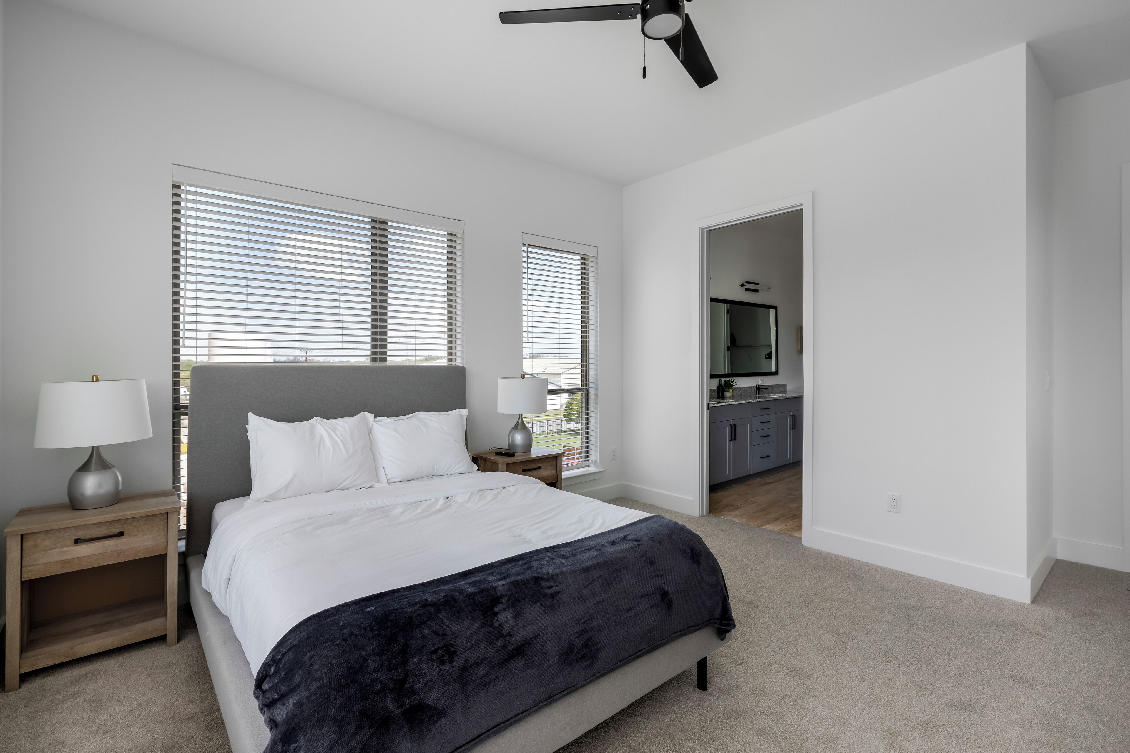 A bedroom with a bed, bedside tables, a ceiling fan, and a view into the kitchen.
