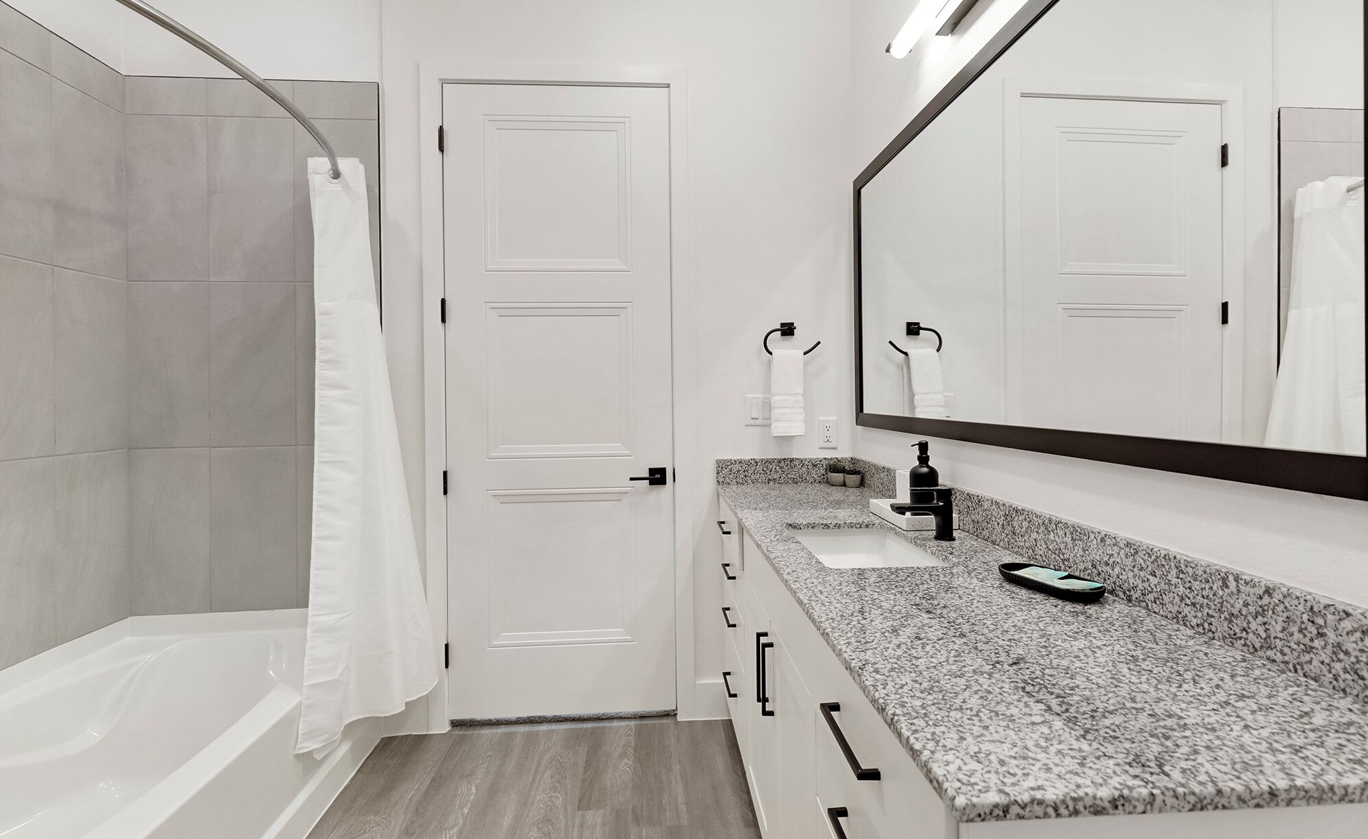 Une salle de bain moderne avec une baignoire, un lavabo doté d’un plan de travail en granit, un grand miroir et une porte.