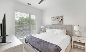 A modern bedroom with a large bed, two bedside tables with lamps, a flat-screen TV, and a window with blinds. - Landing Apartments Denton (Denton)