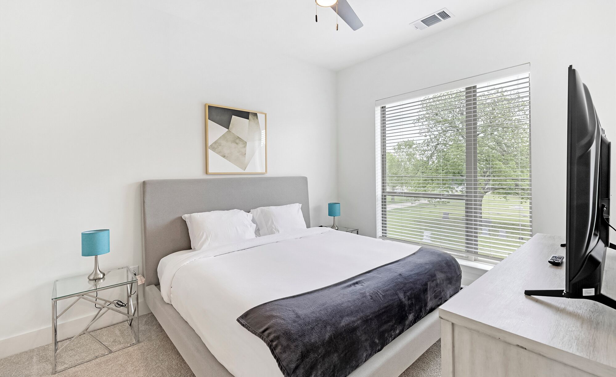 A bedroom with a bed, a TV, a window with blinds, and a painting on the wall.