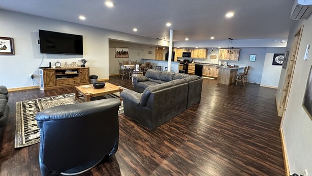 A spacious living area with a large gray sectional sofa, a black armchair, and a wooden coffee table. A flat-screen TV is mounted on the wall above a wooden entertainment unit. The room features dark wood flooring, a patterned rug, and a kitchen area with wooden cabinets and a dining table. - Large, Family-Friendly Lake Home, On Snowmobile/ATV Trails, W/D, Firepit (Harshaw)