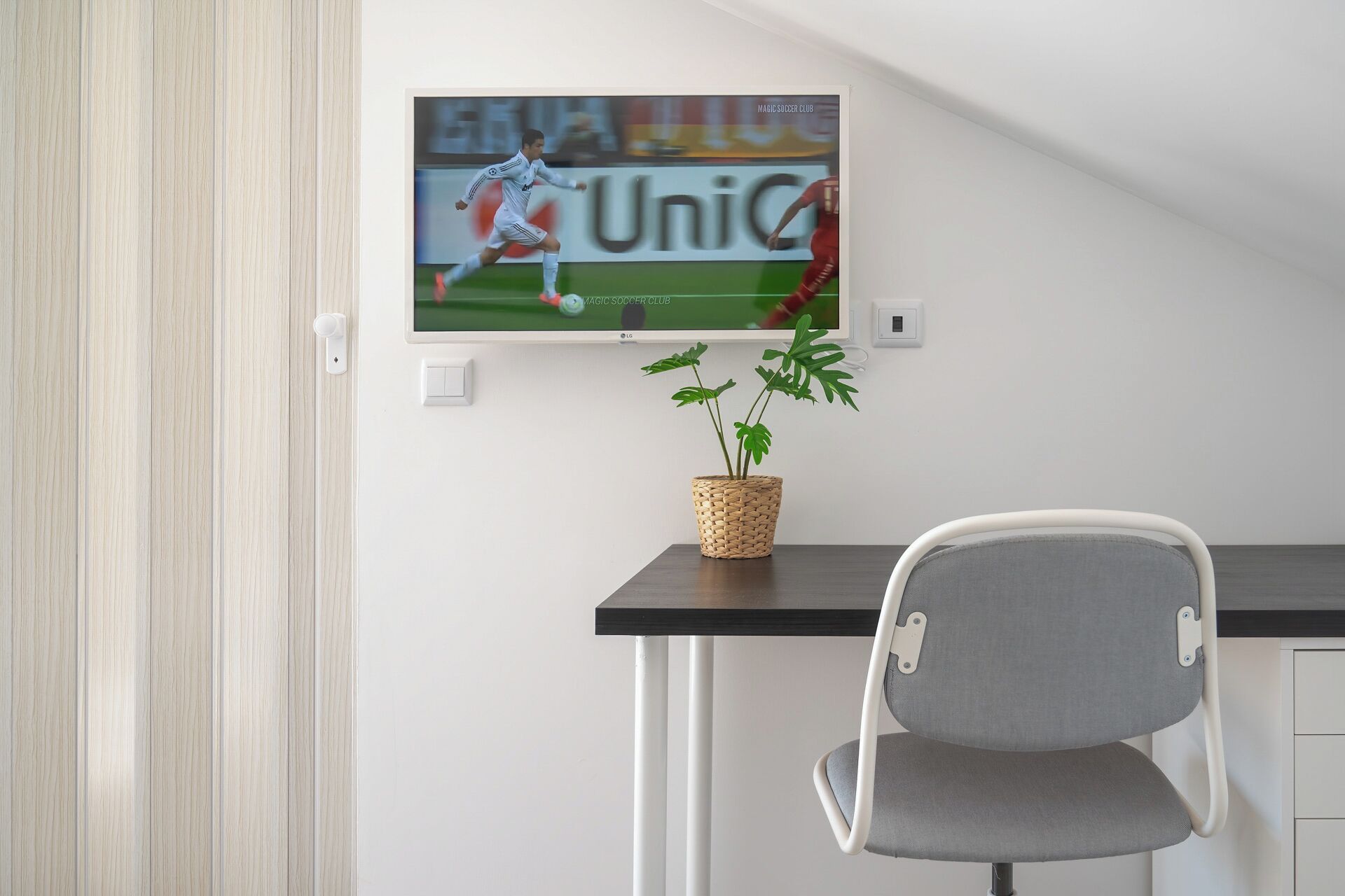 A modern room with a TV mounted on the wall displaying a soccer match, a small potted plant on a table, and a grey chair.