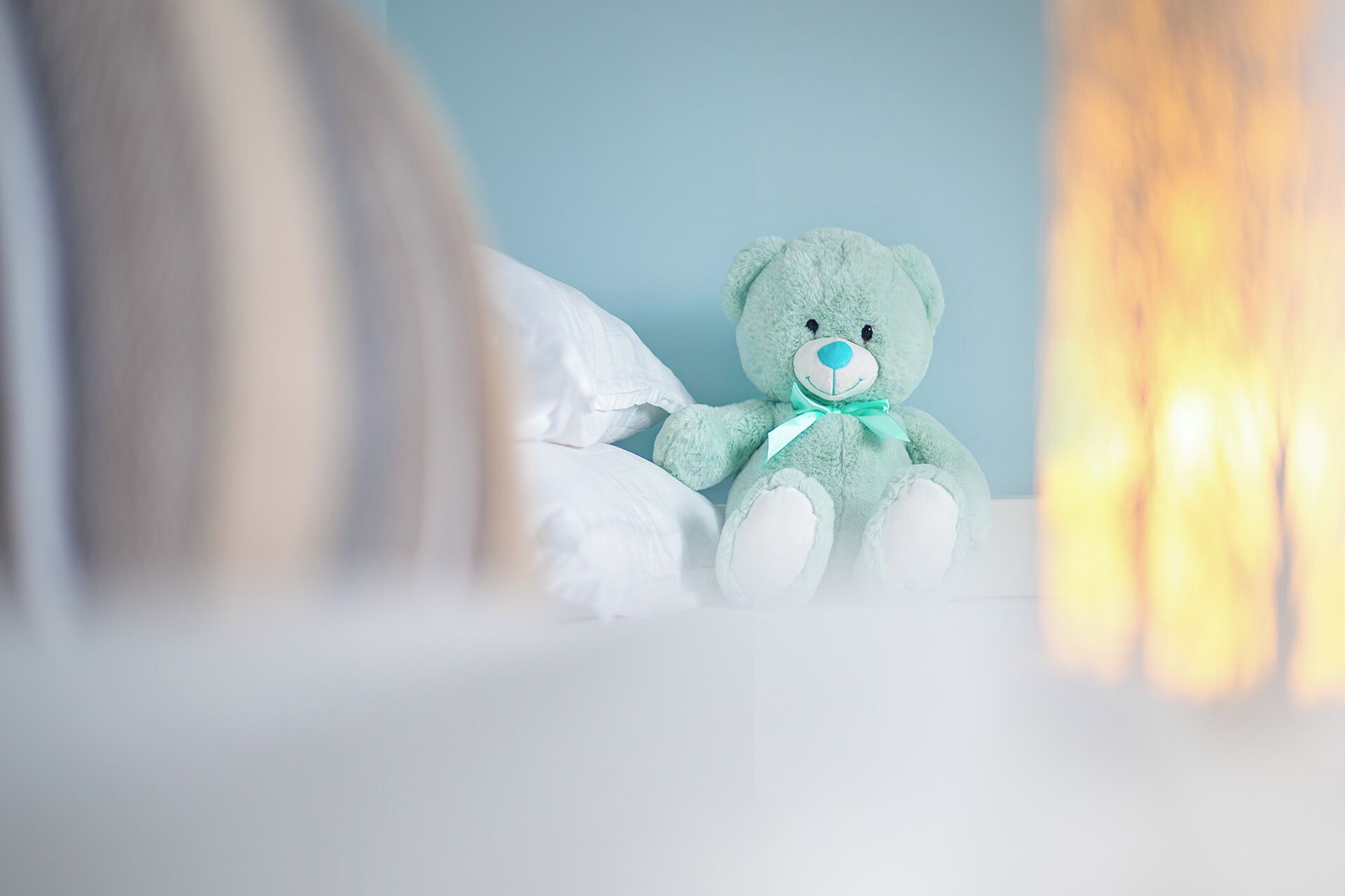 Un osito de peluche verde sobre una cama con almohadas blancas y una pared azul al fondo.