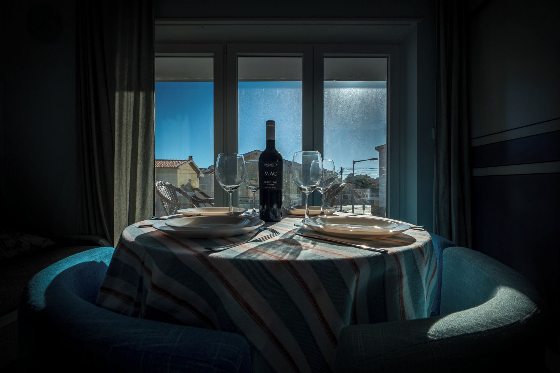Een eettafel met een fles wijn, glazen en borden, voor een raam met uitzicht op gebouwen en een heldere lucht.