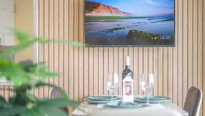 Un comedor con pared de paneles de madera, un televisor de pantalla plana mostrando un paisaje costero, y una mesa dispuesta con platos, vasos y una botella.