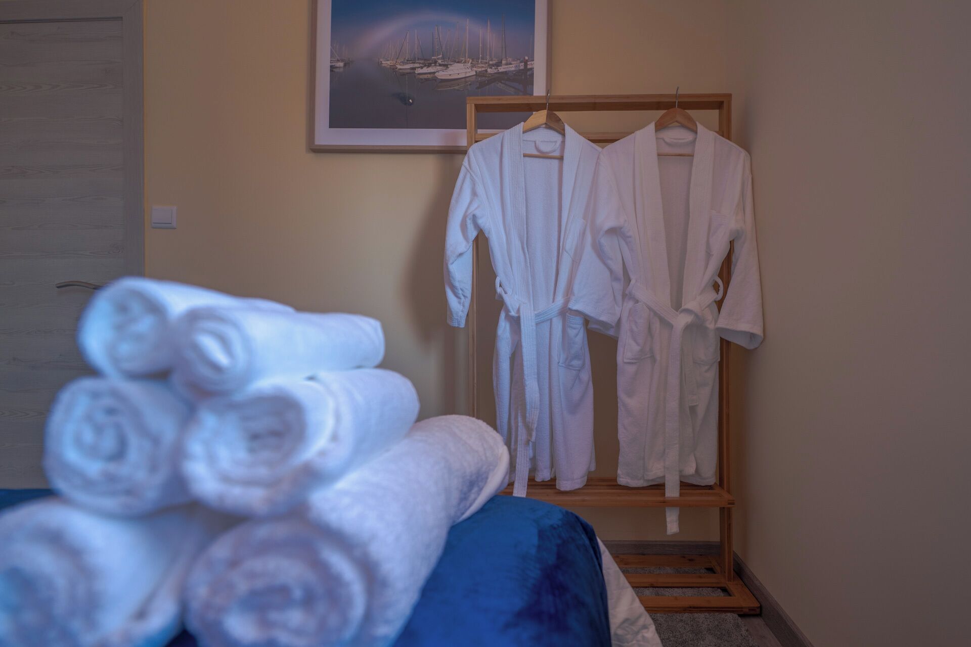 A neatly arranged bathroom with a stack of rolled towels, a wooden rack with white robes, and a framed picture of a marina on the wall.