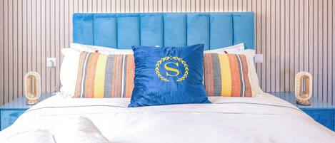Ein ordentlich bezogenes Bett mit blauem Kopfteil, weißer Bettwäsche und einem blauen Kopfkissen mit einer goldenen 'S'.