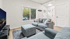 En moderne stue med en grå sofa, et fladskærms-tv og et stort vindue med udsigt til træer og en hvid pickup-truck.
