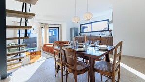 Un coin repas avec une table en bois dressée pour quatre personnes, entourée de chaises en osier. On aperçoit une cuisine à l’arrière-plan, avec une fenêtre au-dessus de l’évier.