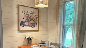 A dining area with a wooden table and benches, a hanging light fixture, a framed artwork on the wall, and a window with curtains.