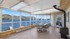 Amplia habitación con ventanas grandes que ofrecen vistas a un lago y colinas. Cuenta con una mesa de comedor, sillas y una chimenea.