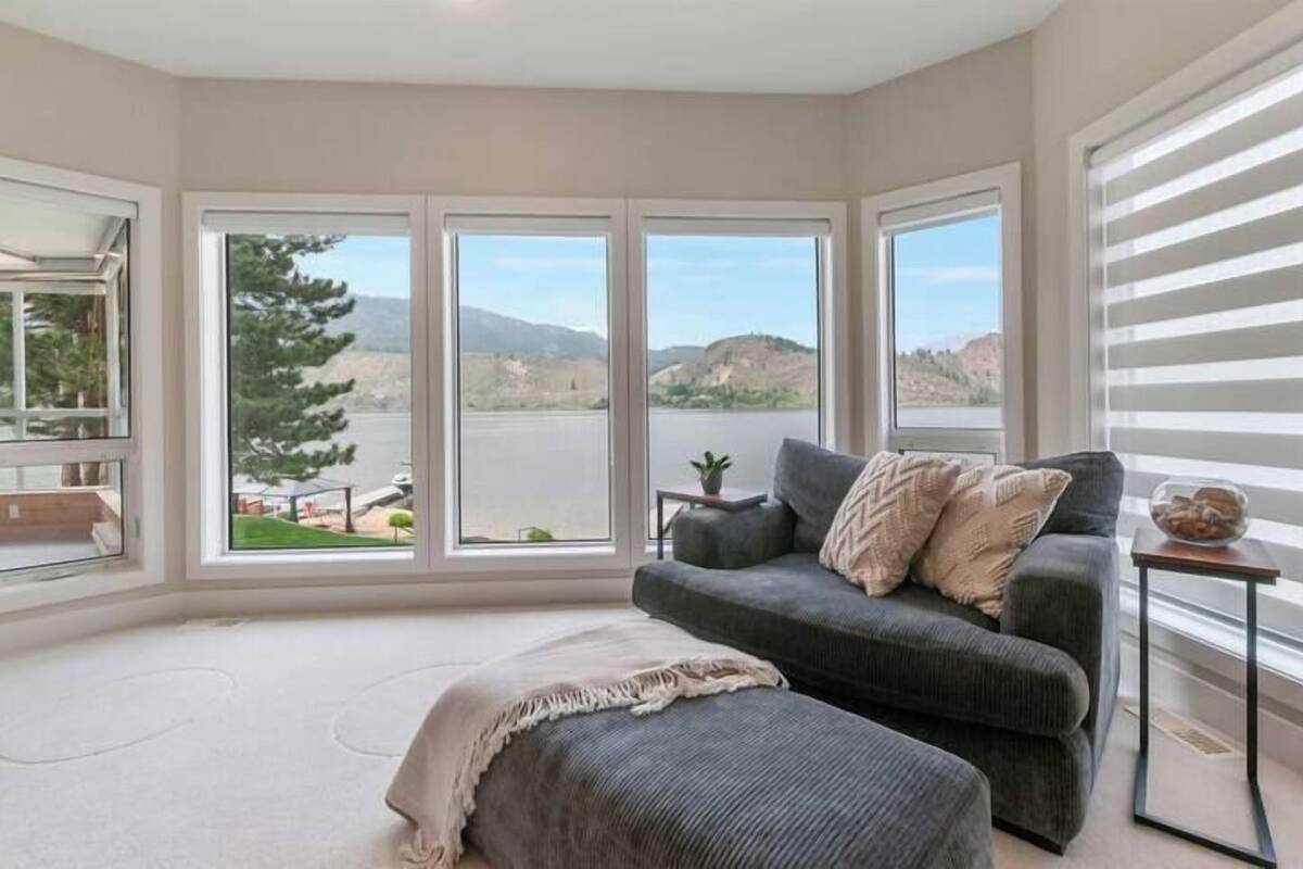 Una sala de estar moderna con un sofá gris, una manta color beige y vistas a un lago y colinas a través de ventanas amplias.