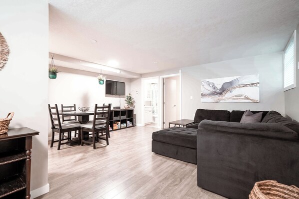 A modern living room with a grey sofa, wooden dining table, and chairs. There is a flat-screen TV mounted on the wall, and a painting above the sofa.