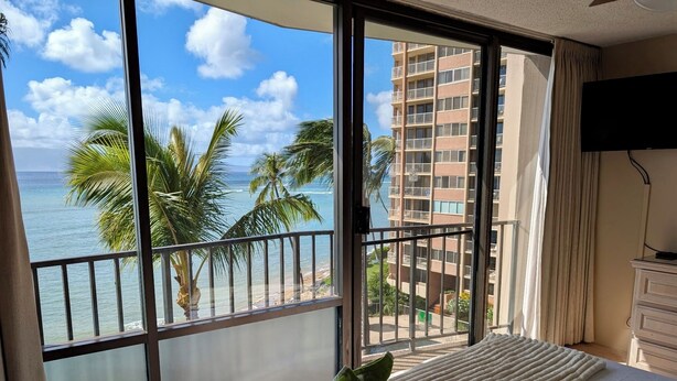 A hotel room with a large window overlooking a beach and palm trees, a bed with a white comforter, and a green and white pillow.