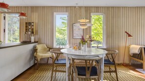 A dining area with a round table, chairs, and a vase with flowers. There are windows with views of greenery and a wall-mounted shelf with books and a lamp. - Cozy home in Vejby with WiFi (Vejby)