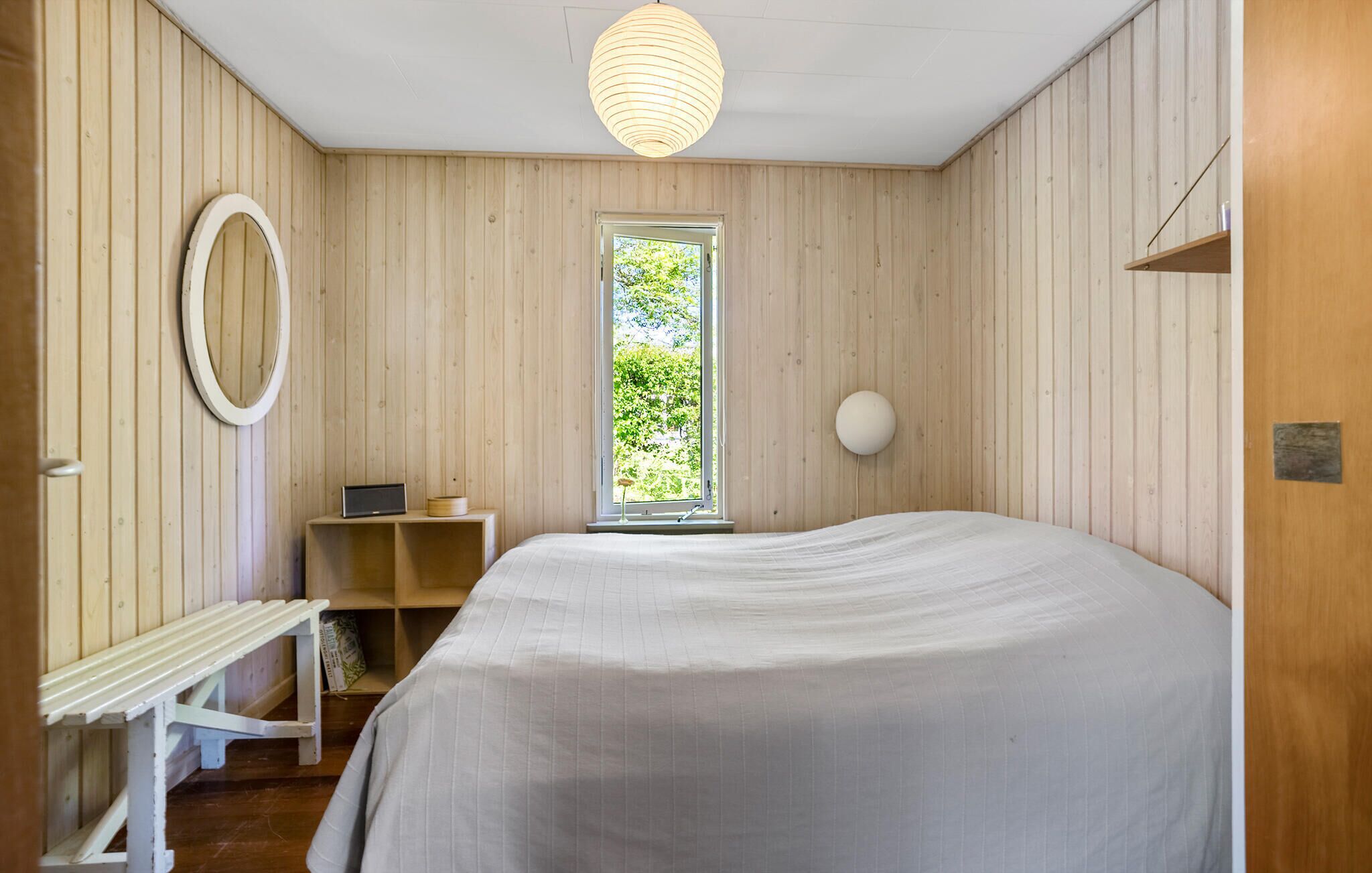 A bedroom with a large bed, a bedside table, a mirror, a window, and a hanging light fixture.