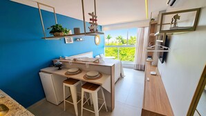 A compact hotel room with a kitchenette, a bed, a window with a view of greenery, and a small dining area. - Porto de Galinhas Center | 3 Min from the Beach (Ipojuca)