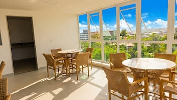 Una habitación con una mesa redonda y sillas, ventanas amplias con vistas a zonas verdes y edificios, y una puerta que da a otra habitación.