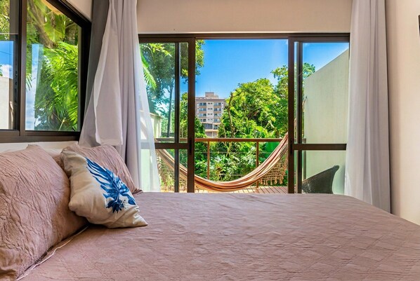 Un dormitorio con una cama grande, vista a una hamaca y un balcón con vegetación.