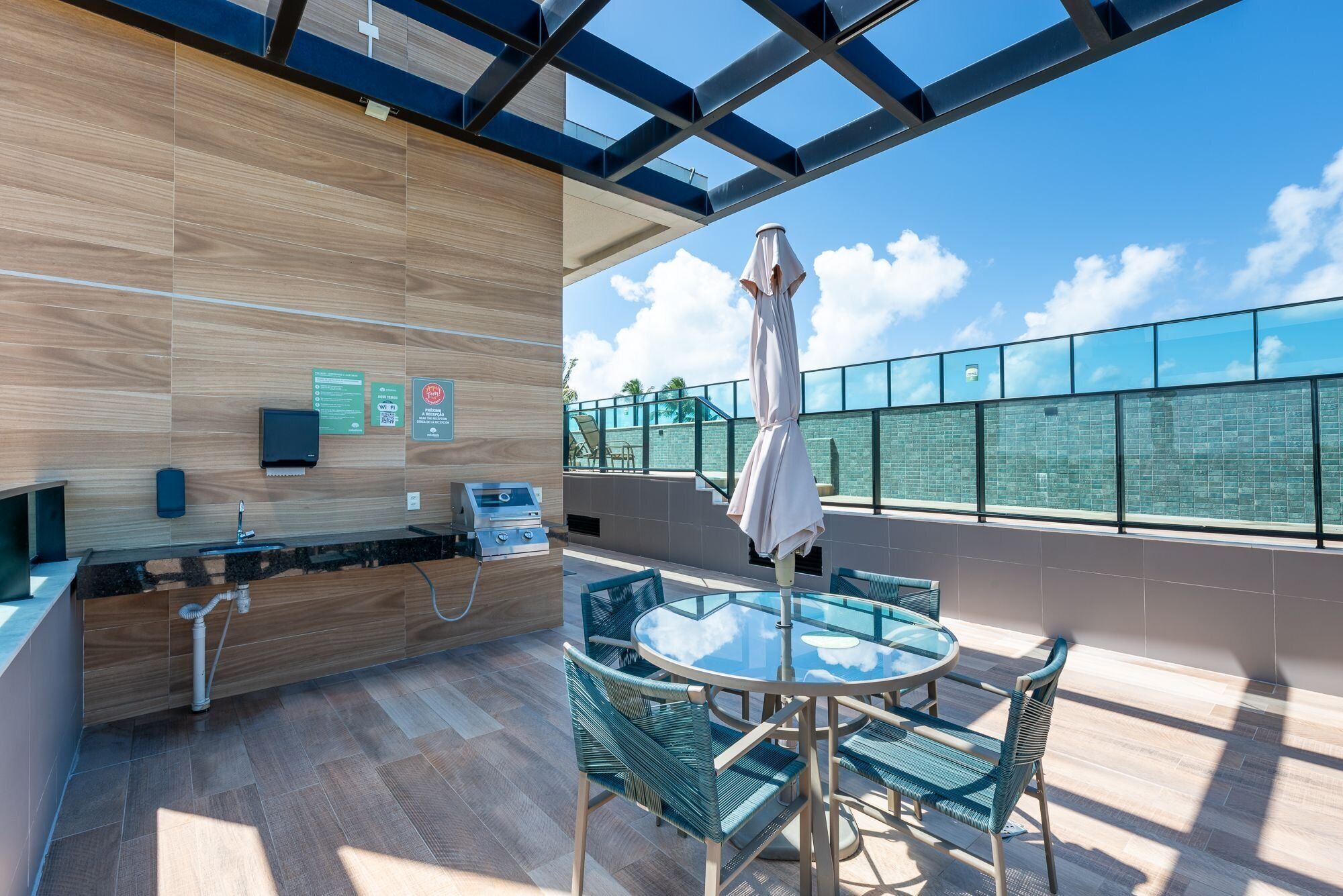 Un espace extérieur moderne avec une table et des chaises en verre, un comptoir en bois doté d’un évier, et une vue sur le ciel et les nuages.