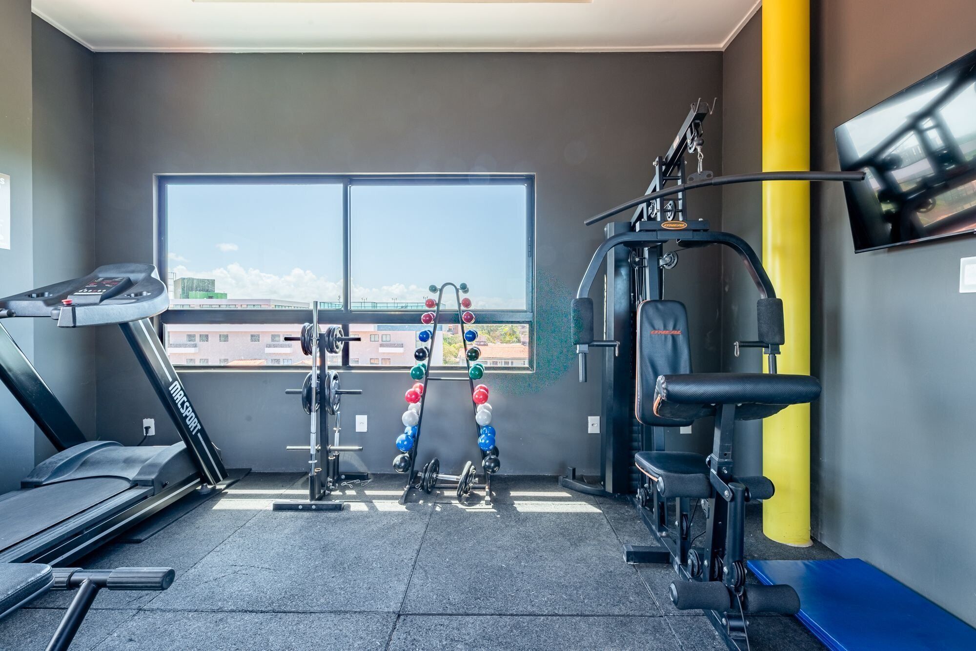 A gym with a treadmill, weightlifting equipment, and a window with a view of buildings.