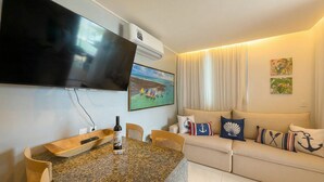 Een woonkamer met een flatscreen televisie, een houten salontafel, een bank met sierkussens met een nautisch thema, en een schilderij van een strandgezicht.