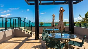 Een balkon met een glazen hekwerk, een ronde glazen tafel en twee beige parasols.