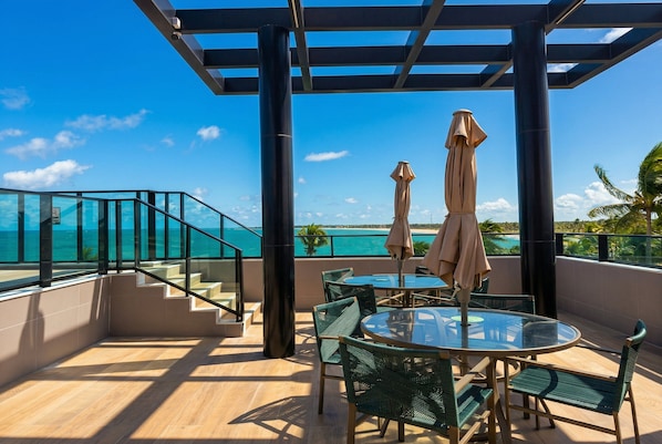 A balcony with a glass railing, a round glass table, and two beige umbrellas.