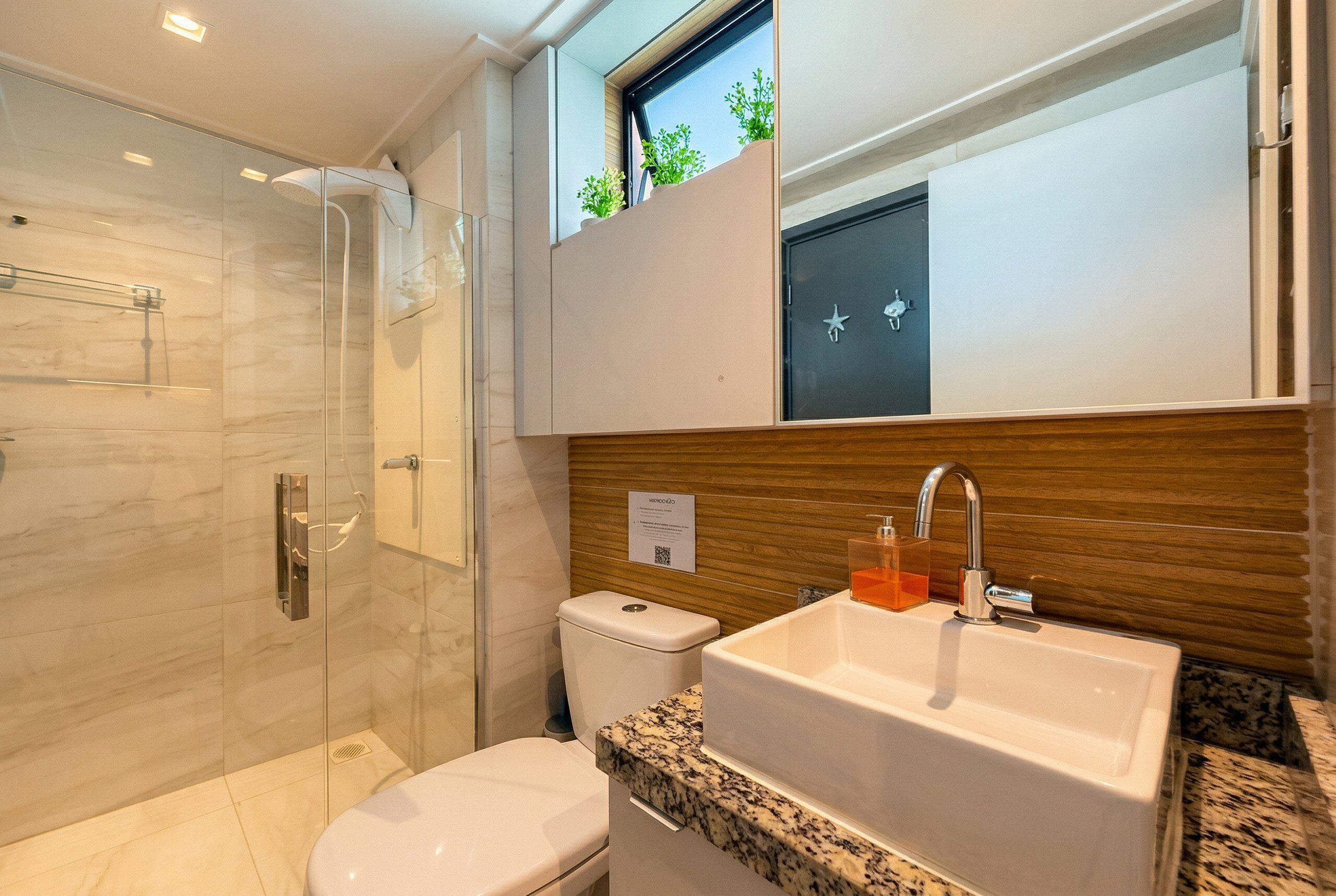 A modern bathroom with a glass shower enclosure, a white toilet, a sink with a wooden countertop, and a window with plants.