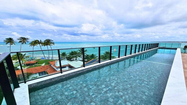 Een lang, smal overloopzwembad met helderblauw water, omgeven door een glazen balustrade. Achtergrond: palmbomen en uitzicht op de oceaan.