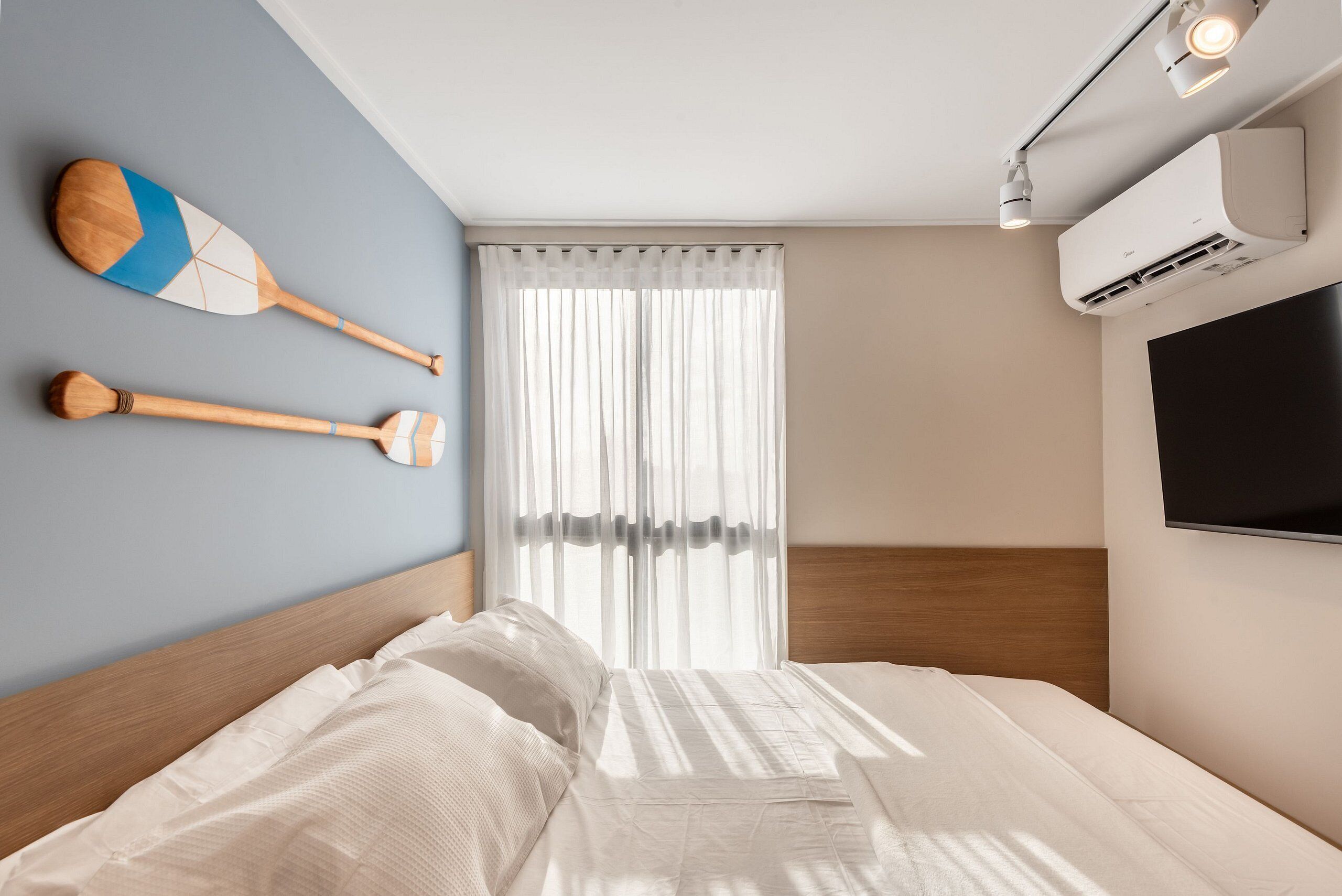 A modern bedroom with a bed, a wall-mounted TV, and a window with sheer curtains.