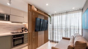 A modern kitchen with a built-in microwave, stove, and oven, a flat-screen TV mounted on a wooden wall, and a seating area with a sofa and curtains. - Flat 301 Makadamia Beachfront (Ipojuca)