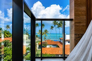 A room with a large glass window offering a view of a beach, palm trees, and ocean. - Beachfront with ocean view (Ipojuca)