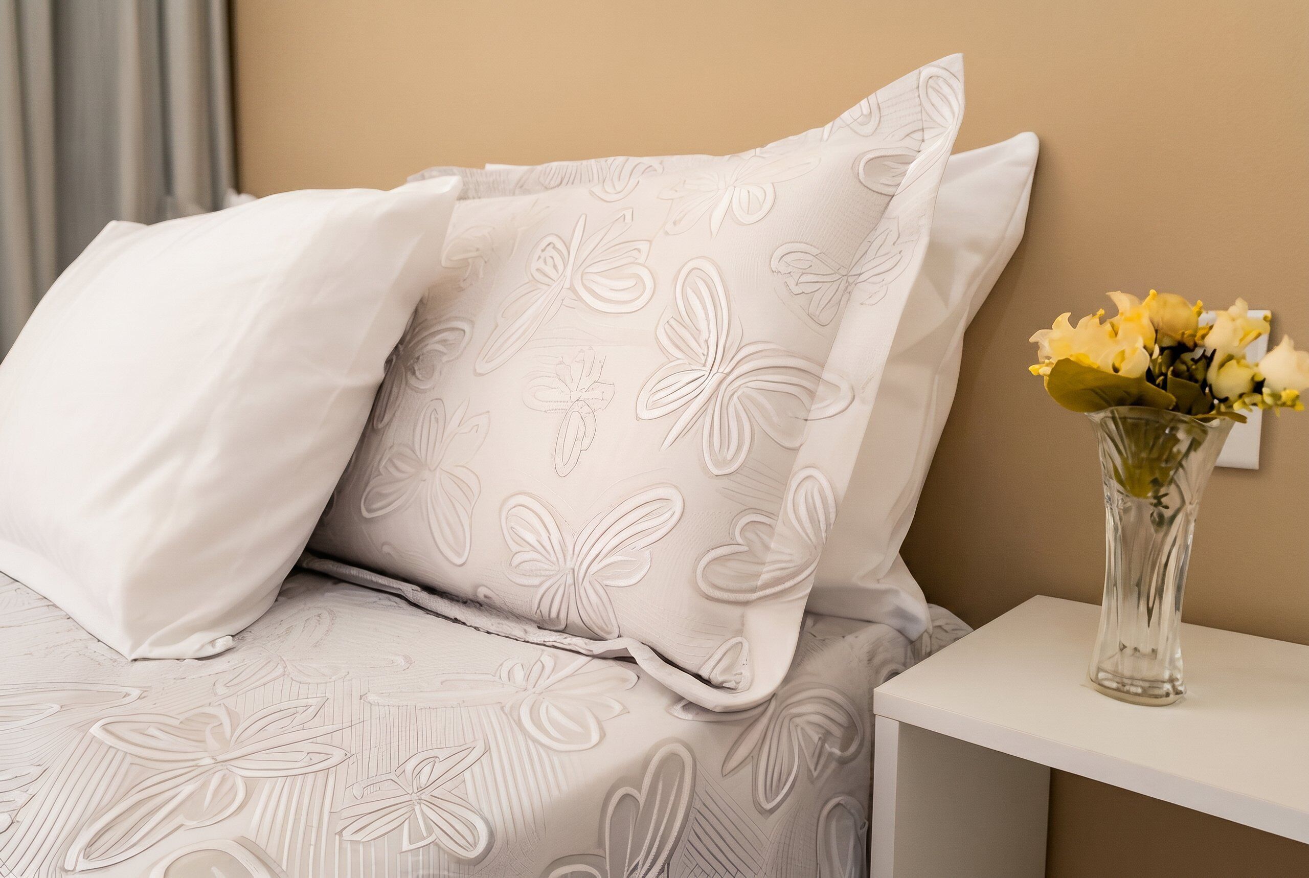 A bed with patterned bedding, a white bedside table, and a vase with flowers.