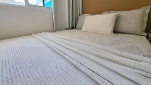 A neatly made bed with a patterned bedspread, a plain pillow, and a headboard against a wall with a window. - 2BR Apartment in Porto de Galinhas | Near Beach (Ipojuca)