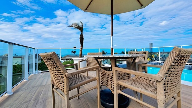 A deck with wicker chairs and a table under an umbrella, overlooking a pool and a city skyline. - 2BR Apartment in Porto de Galinhas | Near Beach (Ipojuca)