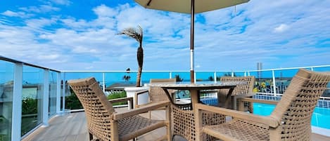 A deck with wicker chairs and a table under an umbrella, overlooking a pool and a city skyline.