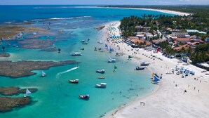 An aerial view of a beach with boats, umbrellas, and buildings. - 2BR Apartment in Porto de Galinhas | Near Beach (Ipojuca)