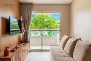 A modern living room with a flat-screen TV, a sofa, a wooden console table, and a sliding glass door leading to a balcony with a view of trees. - Luxury Apartment Near Porto's Natural Pools (Ipojuca)